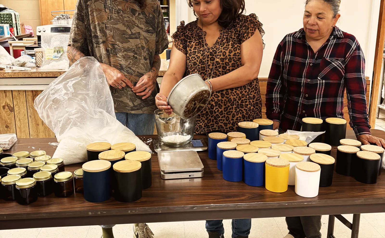 The women measuring wax beside rows of finished candles in amber, black, blue, and cream vessels with wooden lids