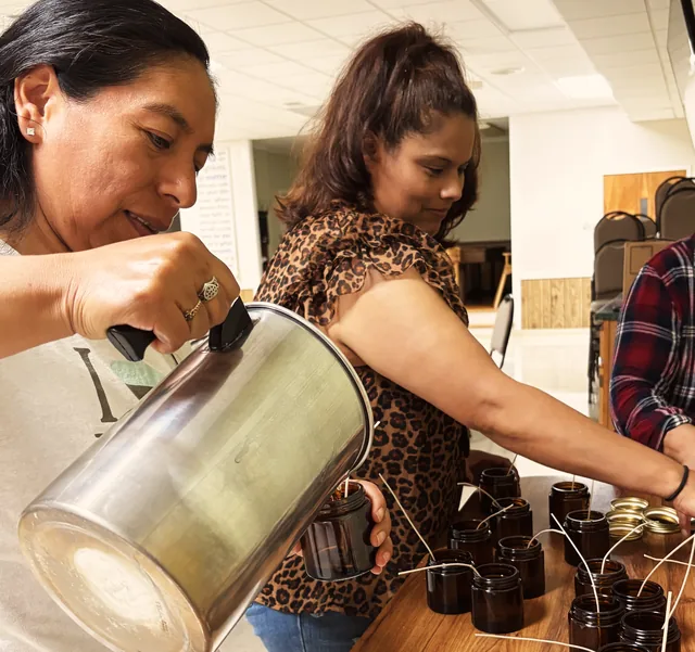 Two women of Las Adelitas pouring warm wax into amber candle jars